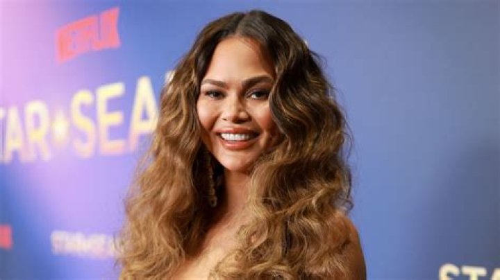 Chrissy Teigen Mimics Cher’s 90s Fitness Video In Sexy Sheer Bodysuit & Curly Wig — Watch